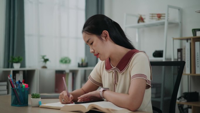 年轻的亚洲女性坐在书桌前，一边在笔记本上写信息，一边在家看书和用手机工作。