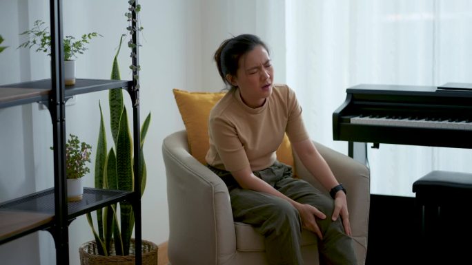 亚洲妇女坐在家里客厅的扶手椅上感到膝盖受伤疼痛。
