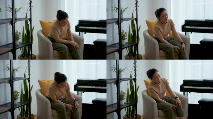 亚洲妇女坐在家里客厅的扶手椅上感到膝盖受伤疼痛。