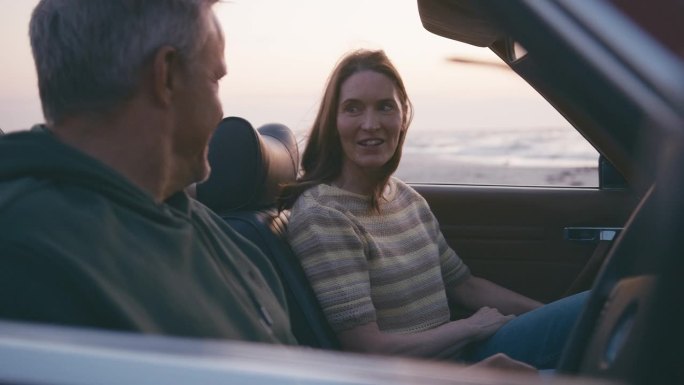 退休的高级夫妇度假坐在经典的敞篷跑车在海滩上观看早晨的日出公路旅行的慢动作拍摄