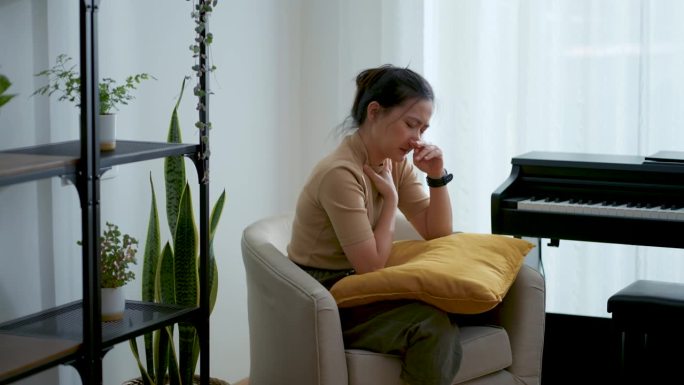 一名亚洲妇女发烧，流鼻涕，鼻塞，喉咙痛，坐在家里客厅的扶手椅上。
