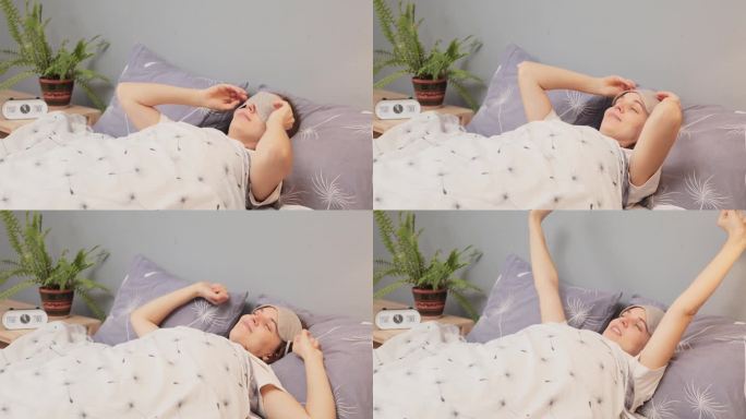 白人女人躺在床上，摘下眼罩，醒来伸伸胳膊，睡个好觉，在舒适的周末早晨休息