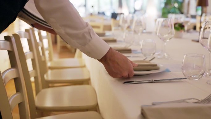 在豪华餐厅的宴会宴会上，制服服务员布置餐桌，为客人准备水晶高脚杯，餐巾纸，白色经典盘子和银质餐具，节