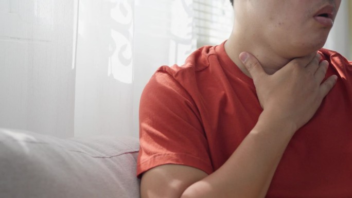 一名亚洲男子因发烧咳嗽，正坐在家里的沙发上，用手抚摸着因感染病毒或感冒而疼痛的脖子。