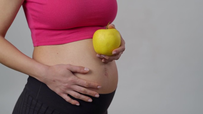特写镜头。一位孕妇站在白色的背景下，一手拿着一个黄色的苹果，另一只手轻轻地抚摸着自己的肚子。