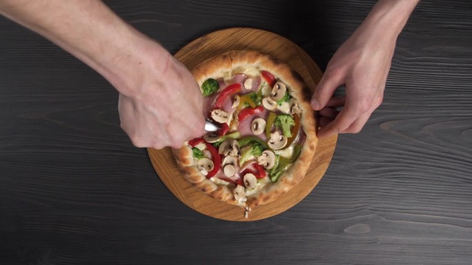 男性的手在木制背景上用披萨刀切披萨。