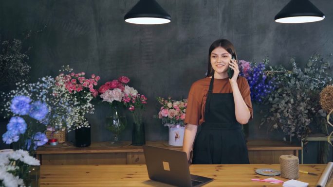 一名女子在被植物和鲜花包围的花店里使用笔记本电脑和手机。卖花者给买花的客户的电话。讨论鲜花的价格。