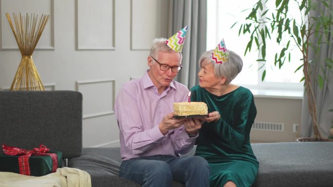 许个愿。家庭高级夫妇戴着派对帽在家里一起庆祝生日纪念日。老人吹灭了生日蛋糕上燃烧的蜡烛。老太太祝丈夫