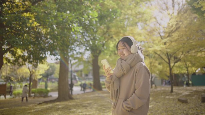 一个寒冷的秋天，年轻的亚洲女子在公园里放松。她用智能手机戴着耳机听音乐。