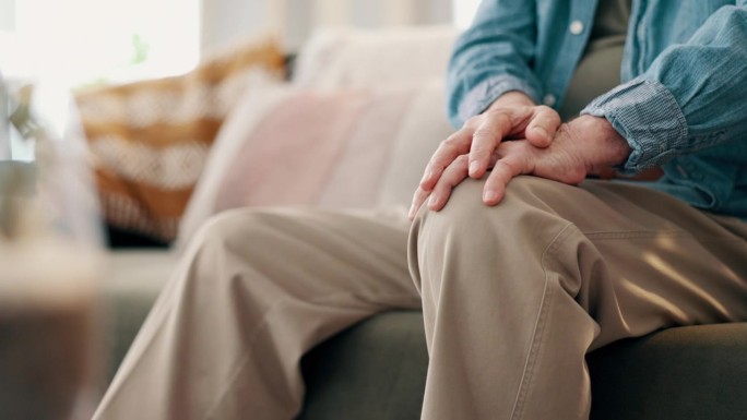 膝盖疼痛，手和老人在沙发上的特写，肌肉，痉挛或炎症在他的家里。Zoom，腿和客厅里患有关节炎、骨质疏