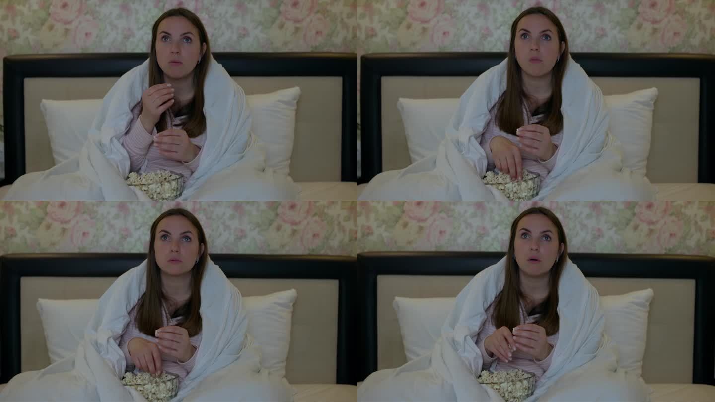 一个女人躺在床上，盖着毯子，拿着爆米花睁大了眼睛，被电视上一个令人惊讶的场景迷住了