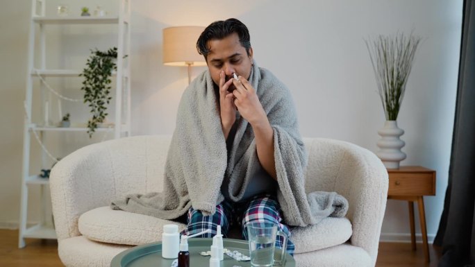 一名印度男子裹在毯子里，坐在沙发上喷鼻喷雾剂