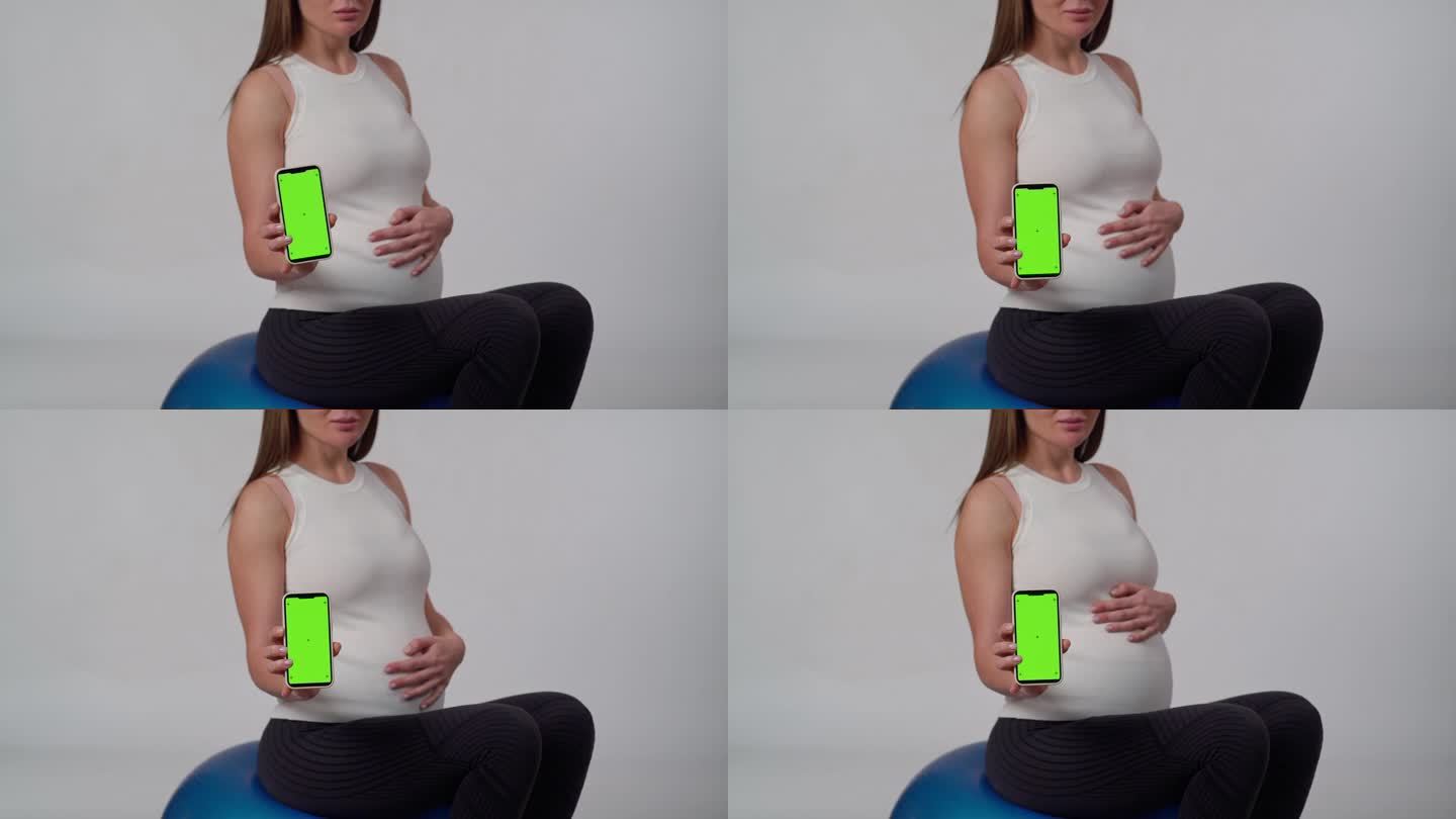 一位无法辨认的孕妇坐在白色背景上，一只手抚摸着肚子，另一只手转动着手机，屏幕正对着摄像头。
