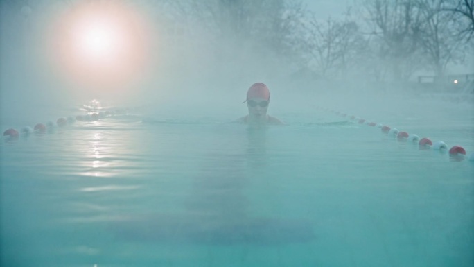 在黎明的大雾天气中，坚定的年轻女子在度假村泳池练习蛙泳