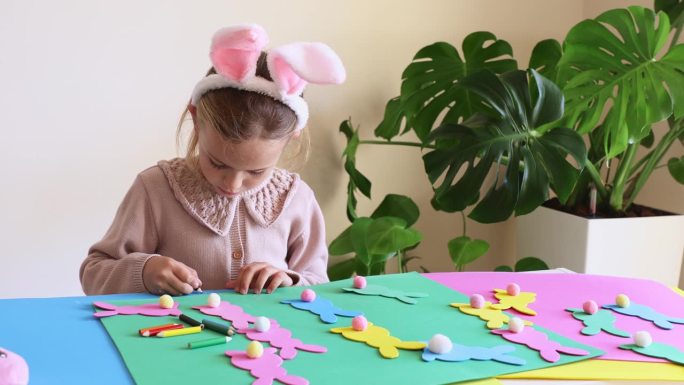 戴着兔子耳朵的小女孩正在用复活节纸制作彩色花环。可爱的儿童工艺品为复活节聚会在家里。节日艺术活动的孩