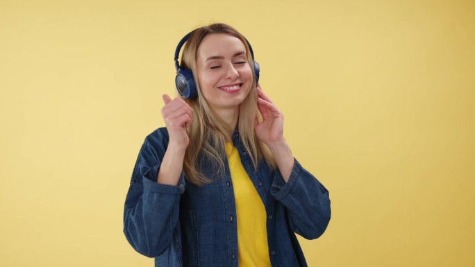 积极的漂亮女人触摸无线耳机和拇指向上的手势在工作室设置。满意的白人女性赞成室内音乐品味。孤立在黄色背