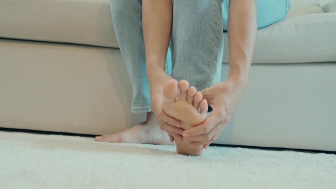 在家坐在沙发上时赤脚疼痛的妇女。足底筋膜炎引起的足部疼痛和长时间清醒。健康医疗理念
