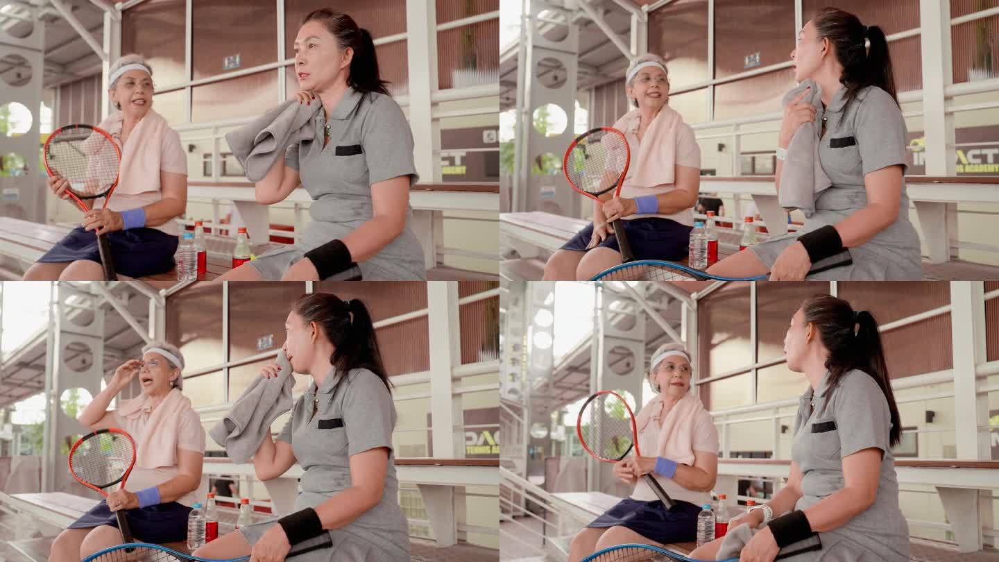快乐健康的亚洲资深网球女选手在网球场边的板凳上休息喝水。在网球场上，资深女子网球选手喝着能量饮料休息