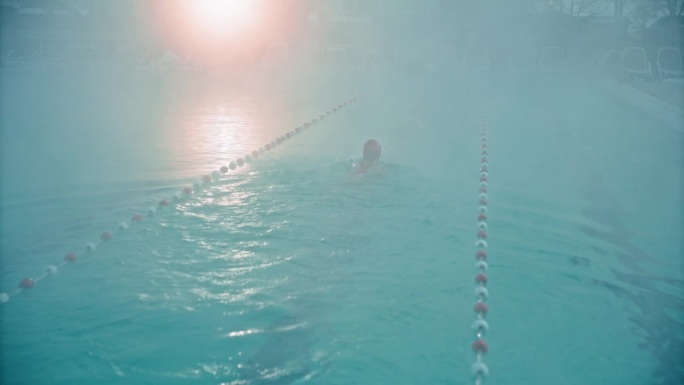 黎明时分，在旅游胜地雾蒙蒙的游泳池里练习自由泳的坚定女子的高角度后视图