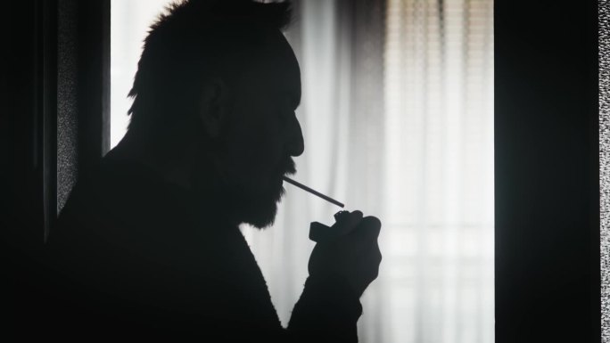 在灯光昏暗的房间里，可以看到一个人的剪影在半透明窗帘的窗户旁抽烟，营造出一种忧郁和大气的效果