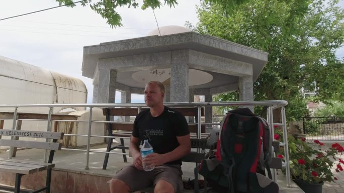 在土耳其的一个旅游景点，一个背包客从背包里拿出一瓶瓶装水，打开它，坐在长椅上喝了一些水。