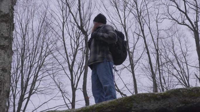 在一个灰暗的冬日，一个穿着法兰绒衬衫、背着黑色背包的男人站在一块巨石的顶端，俯瞰着森林，背景是光秃秃