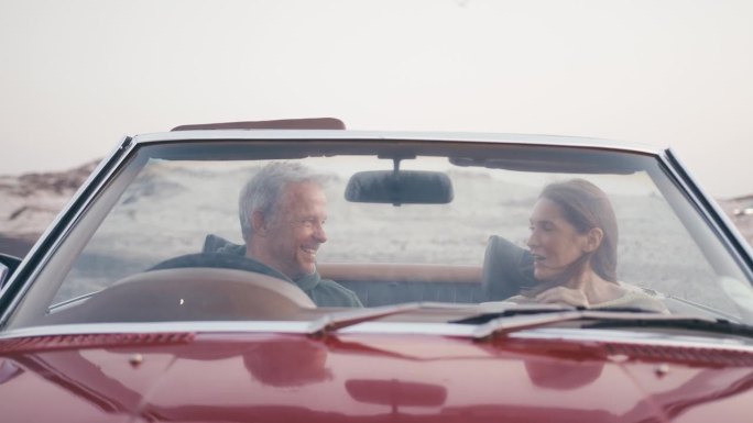 透过挡风玻璃看到退休的高级夫妇在度假，坐在经典的敞篷跑车在海滩上看早晨的日出和喝咖啡的公路旅行-拍摄