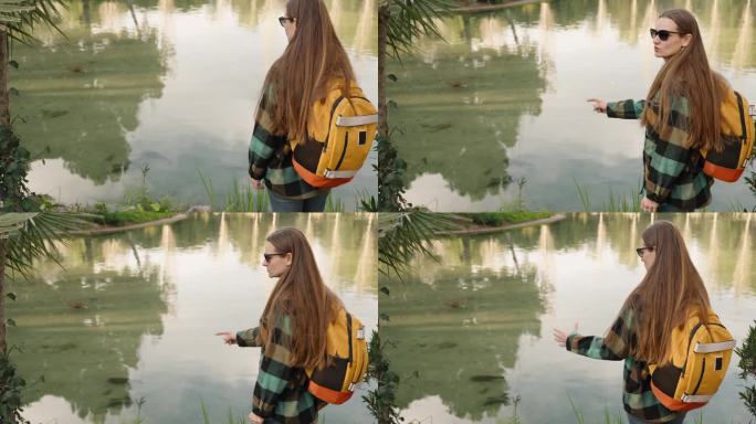 背着背包的女游客观察池塘里的鱼，她转过身来，指着鱼的大小。