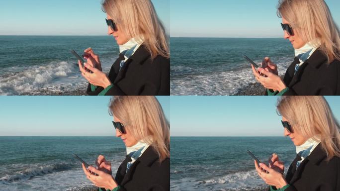 年轻女子在大海或海洋背景下使用智能手机。穿大衣走路。