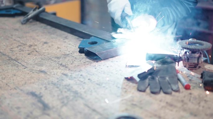 在工厂车间焊接金属板时冒着火花和烟雾，铁匠戴着防护手套和焊接头盔在钢铁材料缝、硬金属制品和制造业工作