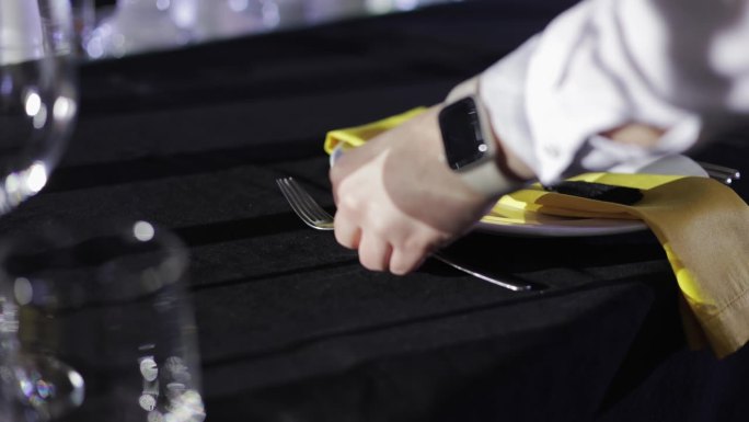 不知名的服务员将两只叉子放在盘子旁边，盘子表面有黄色的纸巾，上面铺着黑色的桌布。专业的餐厅工作人员调