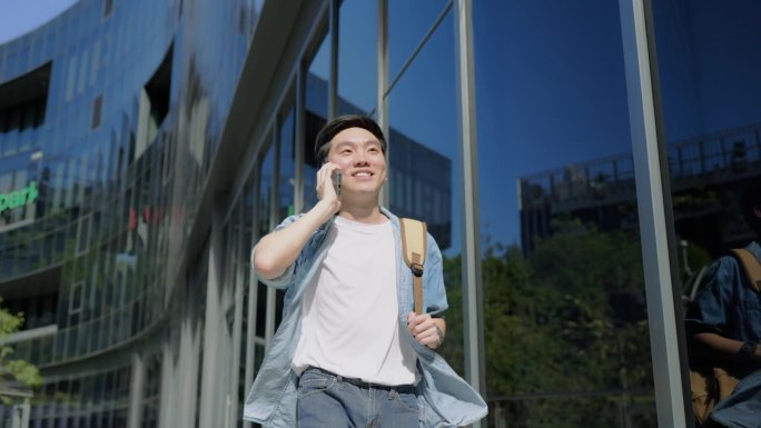帅气的亚洲学生使用智能手机。一个年轻人走在户外，开心地微笑着，用手机聊天
