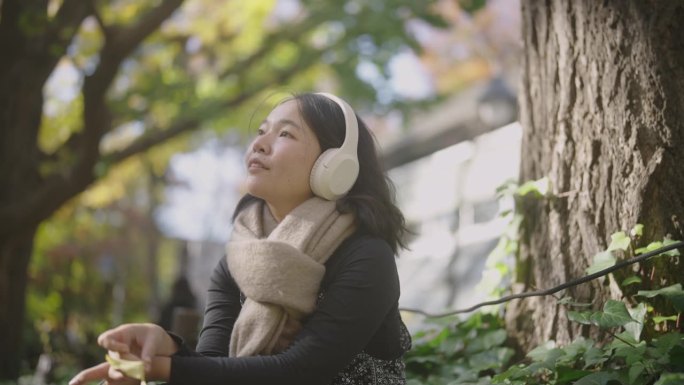 年轻的亚洲女子坐在美丽的风景公园在阳光明媚的日子。她正戴着耳机听音乐。