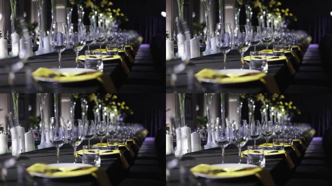 黑色的桌子上装饰着闪闪发光的空酒杯，瓷器拼盘和黄色花朵和餐巾的明亮细节。用餐区旁边的紫色空扶手椅的侧