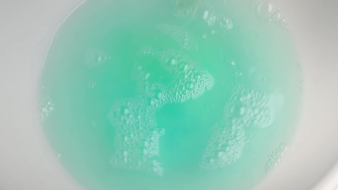 洗衣凝胶胶囊荚与洗衣粉搅拌与棒在热水全帧镜头的蓝绿色液体洗衣粉荚
