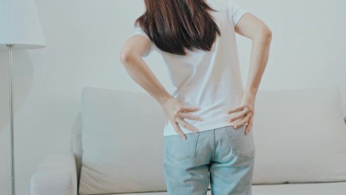坐在家里沙发上时背部疼痛的妇女。成年女性因梨状肌综合征、腰痛和脊柱压迫引起的肌肉疼痛。办公室综合症与
