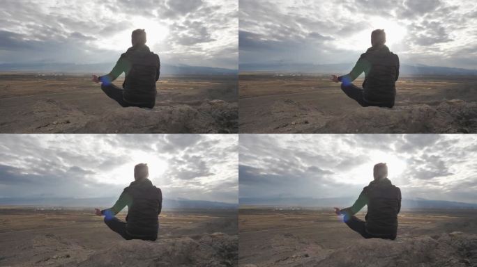 一个扎着马尾辫的年轻人的后景，在日落时分坐在石头上沉思。独自平静地练习瑜伽。精神平衡，与自然和谐相处