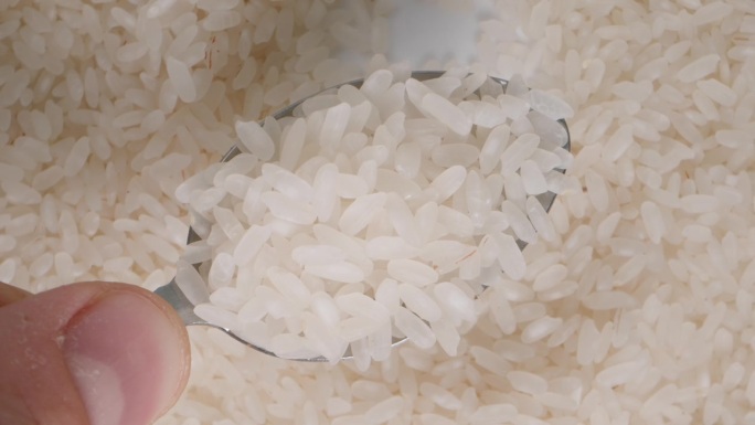 一只手向上举起盛有白米粒的勺子的特写，背景是下面旋转的大米。