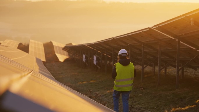 日落时，穿着防护服的女工程师在太阳能发电厂的太阳能电池板中行走的后视图