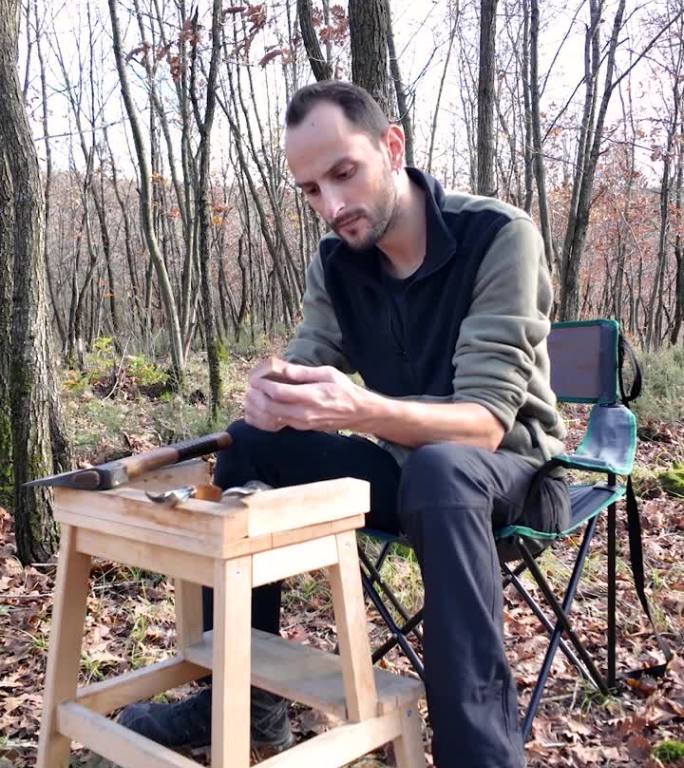 木匠检查了一下雕刻刀的锋利程度，开始磨它。他坐在森林里的露营椅上。