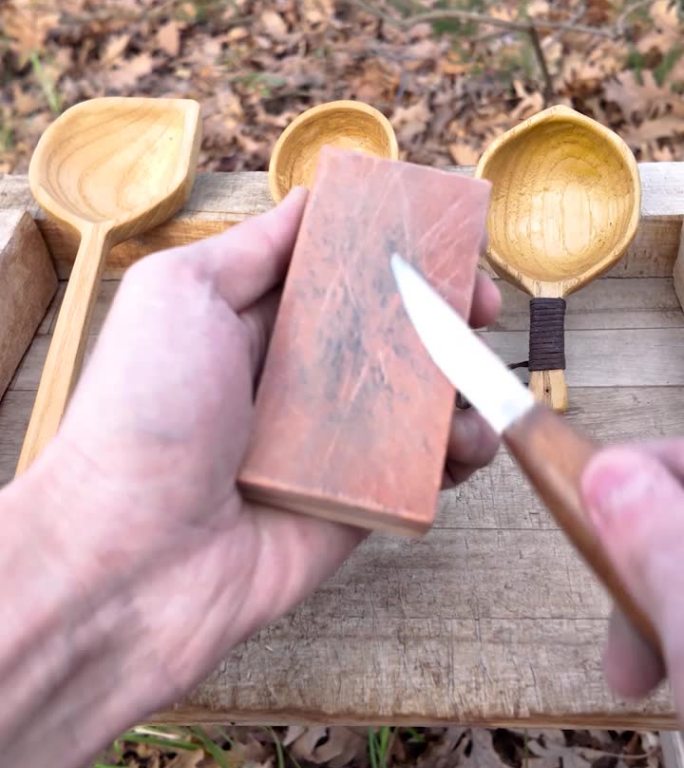 木匠开始磨刻刀的特写。他坐在森林里的露营椅上。