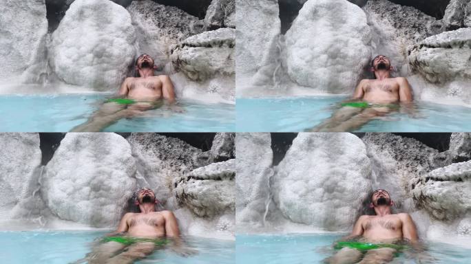 在小瀑布下闭着眼睛享受头部按摩的热硫水浴放松的男人，年轻人倚靠在温泉中的矿物盐石上，从治疗特性中获得