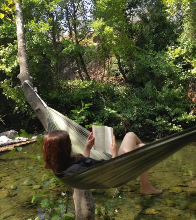 一个女人倚在河边的吊床上看书，吊床系在两棵树之间。营地位于国家公园内，森林茂密。