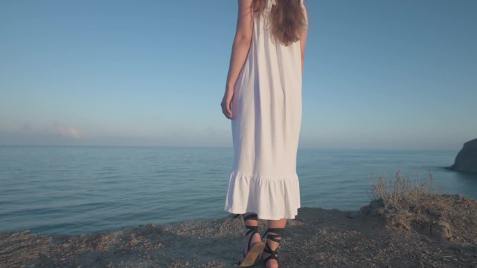 一位身穿白色长裙的年轻金发女子站在海边的棕色岩石峭壁上，欣赏着美丽的海岸景色