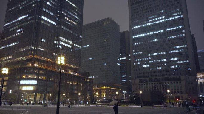 夜晚，东京的摩天大楼上飘着雪花，丸之内的行人打着伞行走