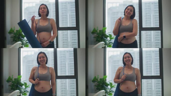 穿着运动服的亚洲孕妇在家里拿着瑜伽垫看着镜头