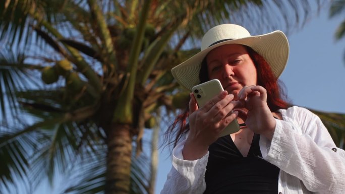女游客站在度假村的椰子树下用智能手机聊天。
