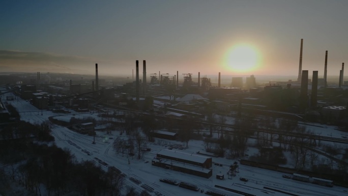 鸟瞰图:一个被雪覆盖的工业区，日落时分，高大、黑暗的烟囱映衬着充满活力的天空