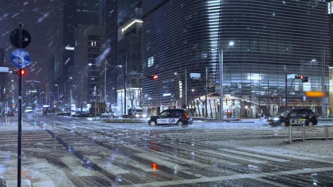 夜晚，东京的摩天大楼上飘着雪花，行人打着伞走着，汽车在东京站的雪道上驶过
