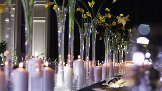 为活动或庆典而精心布置的餐桌。木桌上摆放着多个高大的玻璃花瓶，里面装着黄色的花朵。白色的蜡烛高度不同